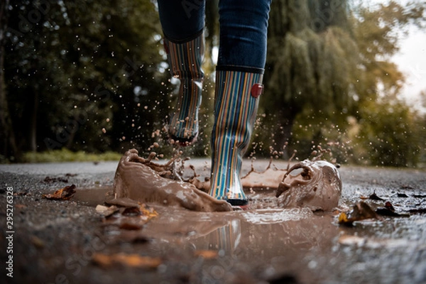 Obraz A woman wearing rubber boots jumps into a puddle on a cold autumn day.