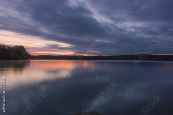 Obraz Landscape; lake and trees before sunrise, long exposure blurred objects, dark clouds and sky, autumn trees and orange sun are reflected in the water