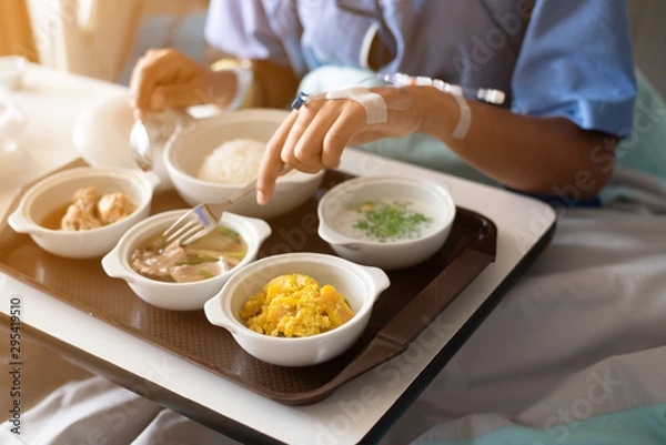 Obraz Woman wearing a blue patient hospital gown, eating food for patients in on the patient bed in the hospital