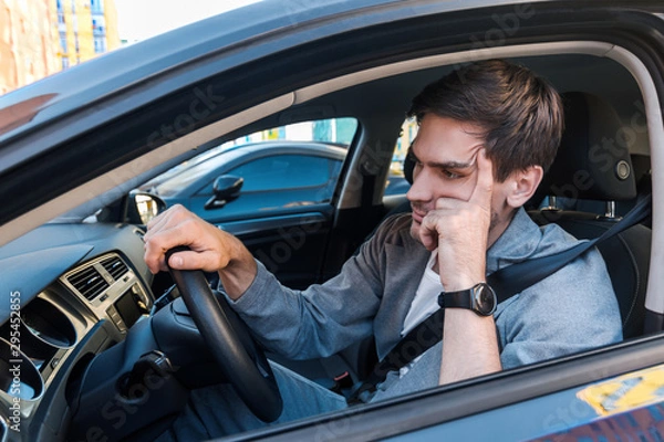 Fototapeta Annoyed tired young man is riding driving car. Businessman is late for meeting. Driver brunette in grey suit stuck in traffic jam. Stressful situations on roads and fast rhythm in modern city.