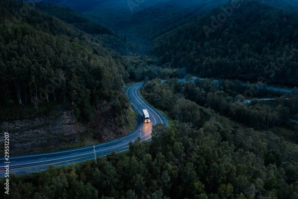 Fototapeta Aerial view of a sharp turn on a mountain road among green forest trees. Semi truck with cargo trailer and bright headlights on a dark highway. P-258 road near Baikal shore in Siberia, Russia
