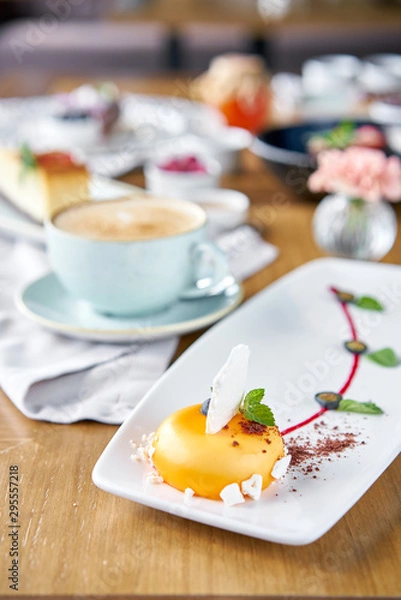 Fototapeta Yellow mousse-cake. Breakfast in the cafe, morning coffee. Cappuccino and lots of desserts on the table. French dessert, blueberry, mint and a piece of meringue on a white plate. Restaurant menu