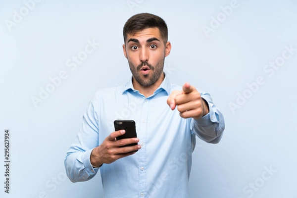 Fototapeta Young handsome man with his mobile over isolated blue wall surprised and pointing front