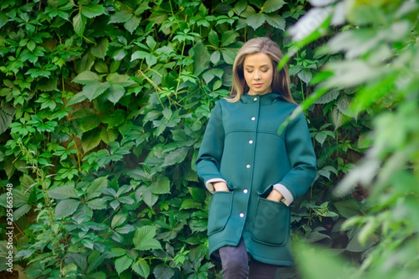 Fototapeta Portrait of a beautiful girl in a green coat on a background of ivy .