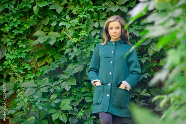 Fototapeta Portrait of a beautiful girl in a green coat on a background of ivy .