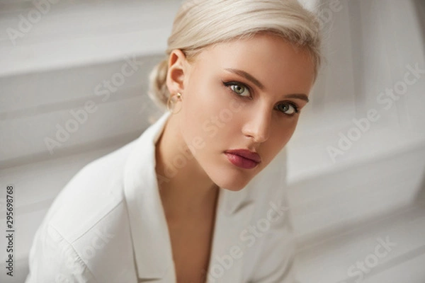 Obraz Modna młoda kobieta o idealnych blond włosach i idealnym modnym makijażu w eleganckim białym garniturze pozuje w studio. Piękna blondynka modelka z czerwonymi pełnymi ustami i głębokimi uwodzicielskimi oczami.