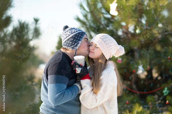 Fototapeta The guy with the girl walk and kiss in the winter forest with a mug of hot drink. A cozy winter walk through the woods with a hot drink. Loving couple, winter holidays