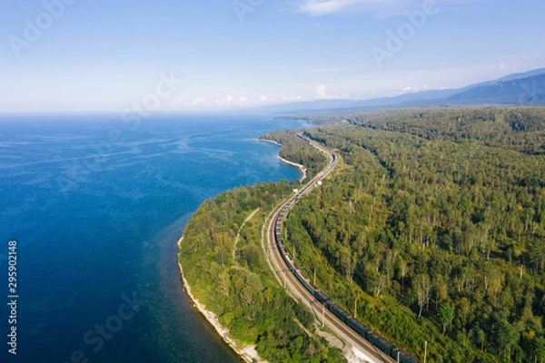 Fototapeta Aerial view of a freight train on the railroad of Trans-Siberian Railway on the shore of Baikal Lake with green forest trees in a sunny summer day. East Siberian Railway in Buryatia, Siberia, Russia