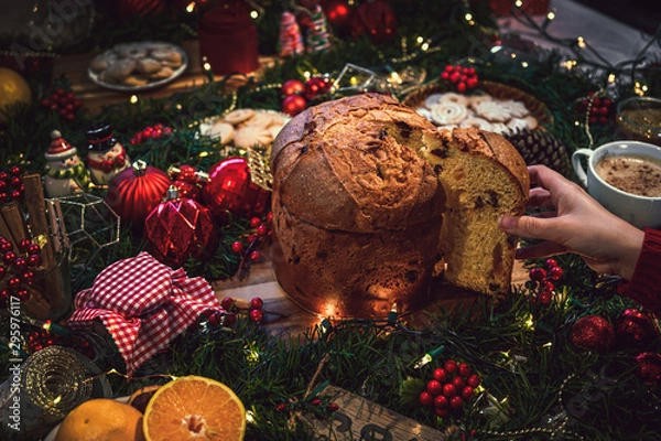 Obraz Young woman holding a slice of panettone at Christmas and in the background a table with holiday season ingredients like hot chocolate, orange, cookies, cinnamon and pine