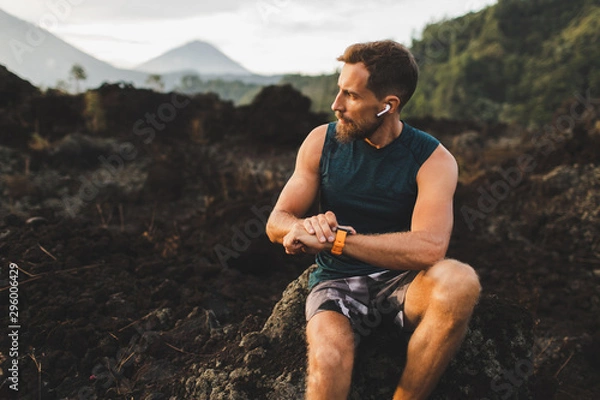 Fototapeta Young hipster runner with beard using smart watch and listening music in wireless headphones before running outdoors.