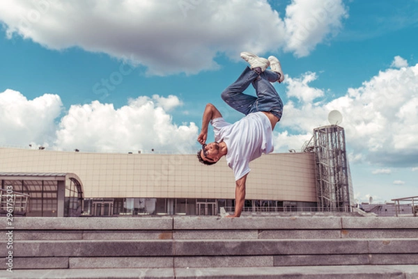 Fototapeta dancer on one arm dances break dance, hip-hop, artist acrobat. Summer city, clouds background. Active youth lifestyle, young male dancer, fitness movement workout breakdancer. Free space for text.