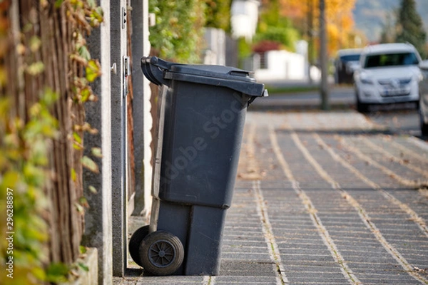Obraz Black garbage bin for residual waste standing on the pavement in a residential area on a sunny autumn day in Germany