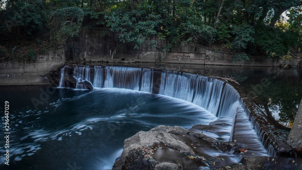 Fototapeta Panoramic View of a Man-Made Waterfall on a River - in a Secluded Spot in the City, Surrounded by Lush Green Trees