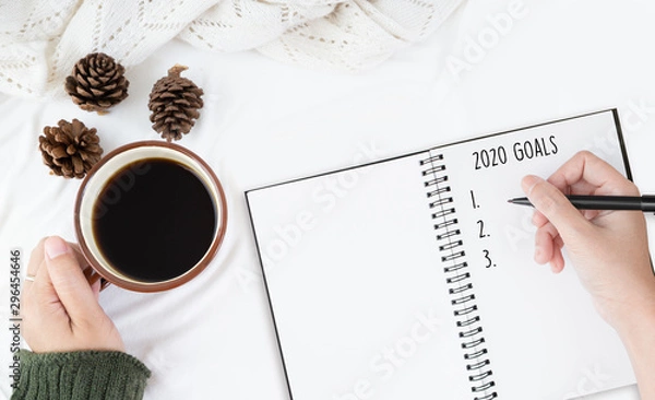 Fototapeta Top view of cup of coffee and pine tree   and dry flower over white table cover and the right hand is writing  down the text for new year goals and resolutions list on white paper note.