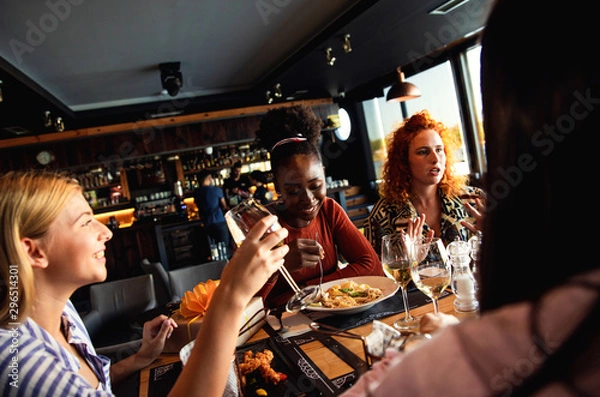 Fototapeta Group of young female friends having fun in restaurant, talking and laughing while dining at table.