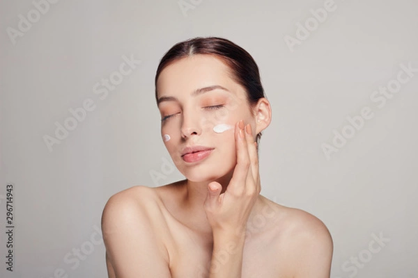 Obraz Young beautiful woman with closed eyes is applying cosmetic cream on her face. Cream on the cheeks.Skin care. Dry skin. Beauty concept.