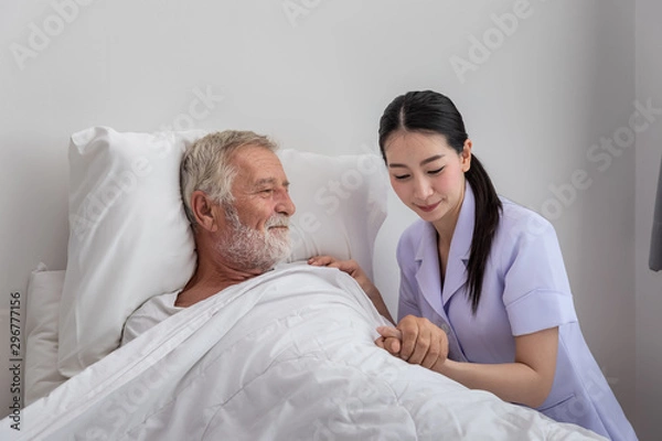 Fototapeta Happy nurse taking care elderly man with blanket in bedroom at nursing home