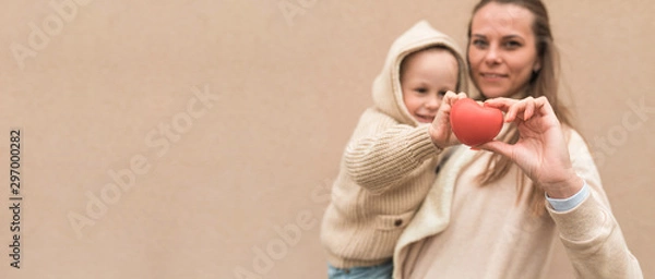 Fototapeta Mom woman with a little boy son 4-5 years old, background wall. In the hands of a toy red heart, concept give life and love, donation. Mother and St. Valentine's Day. Free space for text.