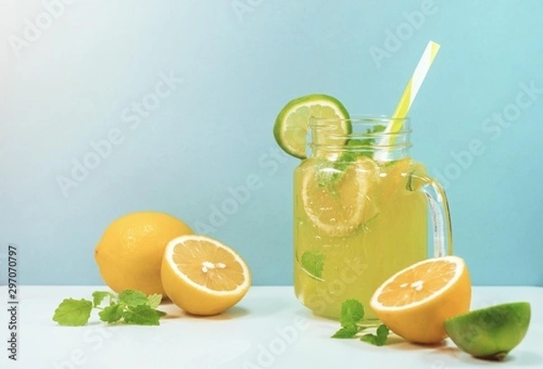 Fototapeta Lemonade drink of soda water, citrus and mint on the light blue background. Cocktail glass with handle and straw, sliced lemon and lime, front view