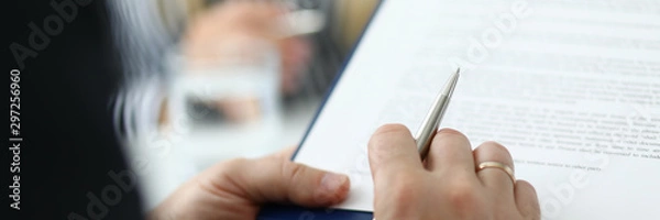 Obraz Focus on male hand holding pen and paper folder with contract. Partner checking conditions of important biz bargain in meeting room. Blurred background