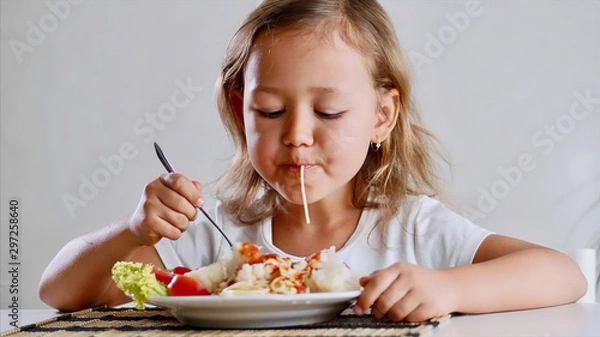 Fototapeta A little girl is eating spaghetti with vegetables at the table in the light room with a fork . Portrait of a kid eat at home in front of the camera.