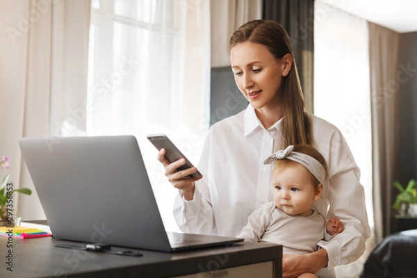 Fototapeta Woman holding smartphone in hand. Little girl sitting on mother's knees and watching cartoons online at laptop. Female sending messages and commenting in social networks, looking at screen of mobile