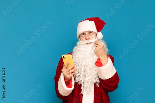 Fototapeta Portrait of a pensive Santa Claus using a smartphone, twirling his mustache and looking thoughtfully upwards against a blue background. Guy in santa suit and smartphone in hand is isolated. New Year