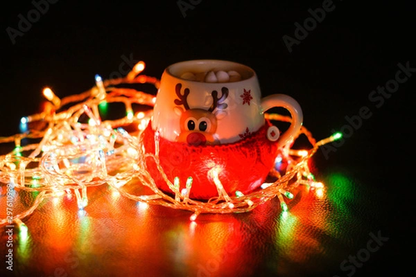 Fototapeta The mug is wrapped in a wool sock. A mug of cocoa and marshmallows, a garland lit next to it. Lights in the dark