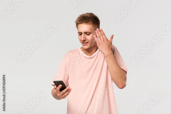 Fototapeta Confused blond teenager in a pink tee looking at the screen of smartphone isolated over light grey background.