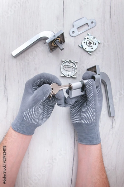 Fototapeta The guy holds in his hands in gray gloves the core of the lock and door keys on a light background. Repair in the house. Installing a new lock on the door. Spare parts for the door on a gray wooden ta