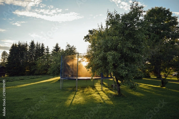 Fototapeta Open children's jump trampoline in the sun on green grass in the yard of finnish house with trees, summer evening