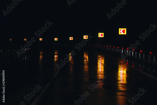 Fototapeta Dark night road with warning signs and bright reflexes on wet asphalt. Driving in rainy weather conditions with bad visibility. Dangerous turn ahead on highway