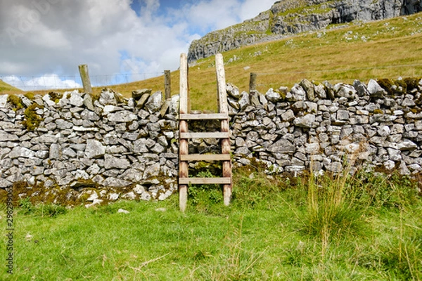 Obraz Detailed view of a wooden, public right of way structure to climb over a stone wall. An empty road leads to the summit of the mountain.