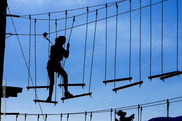 Fototapeta Silhouette of young girl in helmet climbing on high rope course against blue sky. Wooden bars tied to ropes, part of obstacle course in adventure park