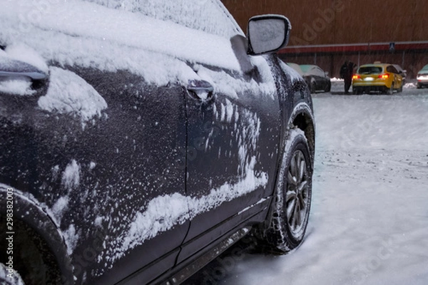 Fototapeta The car is covered with layers of snow and ice. Side view on doors and wheels. Close-up. Taxi driver removes snow from a car in the background. It's snowing, a blizzard. Winter night