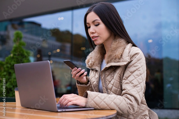 Fototapeta Young beautiful confident woman in down jacket working on laptop with cellphone on street