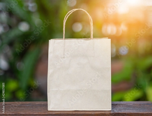 Fototapeta Brown recyclable paper shopping bag Placed on a wooden table, the backdroup is a green tree. For the background image used to campaign, do not use plastic bags.