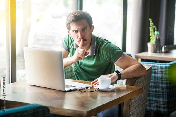 Fototapeta Shhh, Be quiet! Young angry businessman in green t-shirt sitting and working on laptop, looking at camera with finger on lips and command to be silent. indoor shot near big window at daytime.