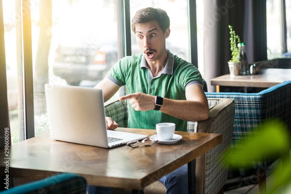 Fototapeta Young shocked businessman in green t-shirt sitting, working, looking and pointing at laptop display with amazed unbelievable face. business and freelancing concept. indoor shot near window at daytime.
