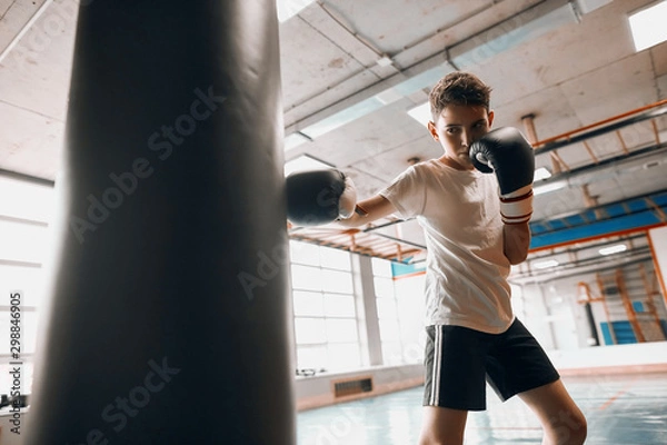 Fototapeta little young hardworking boxer learning to blow the punching bag at sport center, close up photo, effective training process, kid taking up a new hobby