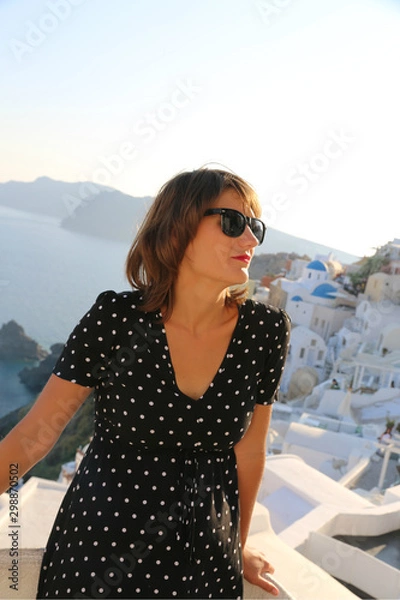Obraz Stylish woman in long black dress standing in luxury hotel up happy in Oia, Santorini, Greece island. Stylish girl tourist in long dress in Santorini Greece.