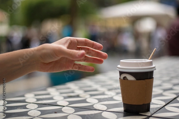 Fototapeta A hand reaches for dark vending coffee in disposable kraft paper cup and plastic lid on table at street cafe.