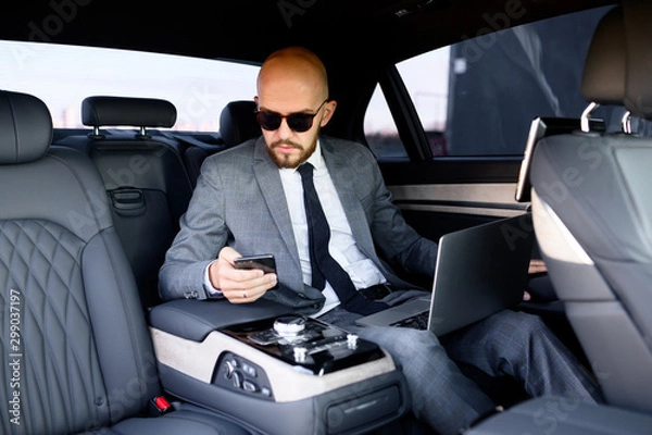 Obraz handsome businessman using his mobile phone in a modern car with a driver in center of the city. Concept of business, success, traveling, luxury 