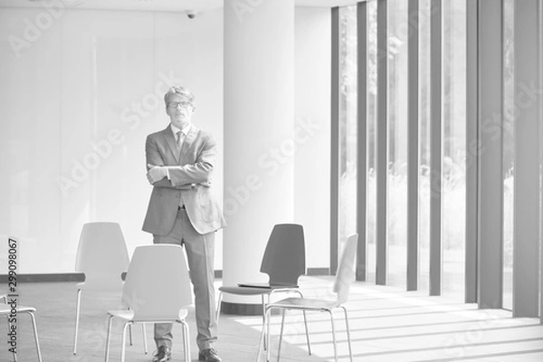 Fototapeta Black and white photo of Confident mature businessman standing with arms crossed amidst chairs in new office