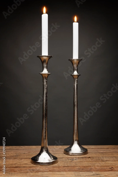 Fototapeta 2 tall silver candle holders and glowing candles, shot on a wooden table