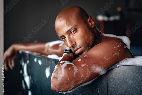 Fototapeta Close up of a sexy Arabic male having a bath in the black stylish tub in thick foam near the window, the man is looking intensely; luxury concept.