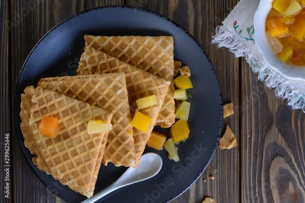 Fototapeta Thin waffles folded as a corner with cream, syrup and slices of fruit on a dark ceramic plate on a wooden table ready for breakfast, top view