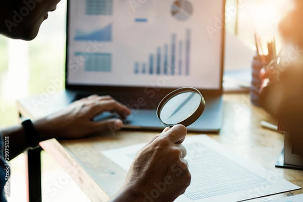 Fototapeta Businessman looking through a magnifying glass to documents. Business assessment and audit. Magnifying glass on a financial report. Concept of search.
