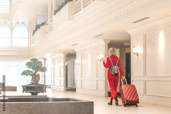 Obraz Full-length portrait of gorgeous lady with blonde hair wearing red jumpsuit entering in the hotel lobby with a backpack and a suitcase. Business trip concept. Horizontal shot. Back view
