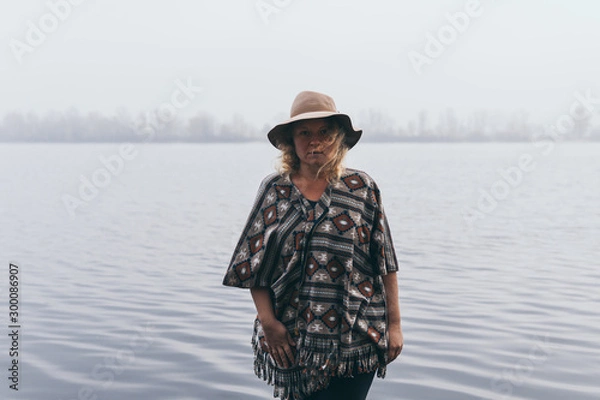 Fototapeta Woman in hat and poncho standing on the river bank and looking towards autumn forest covered with morning fog